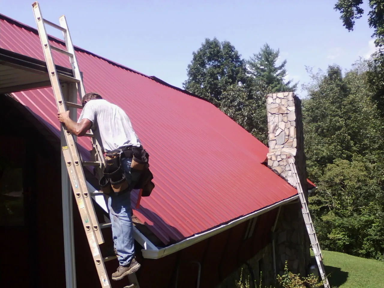 Roofer on a ladder beside a red metal roof and stone chimney — Soffit & Fascia Repair by Roofing Brothers in Broad Top